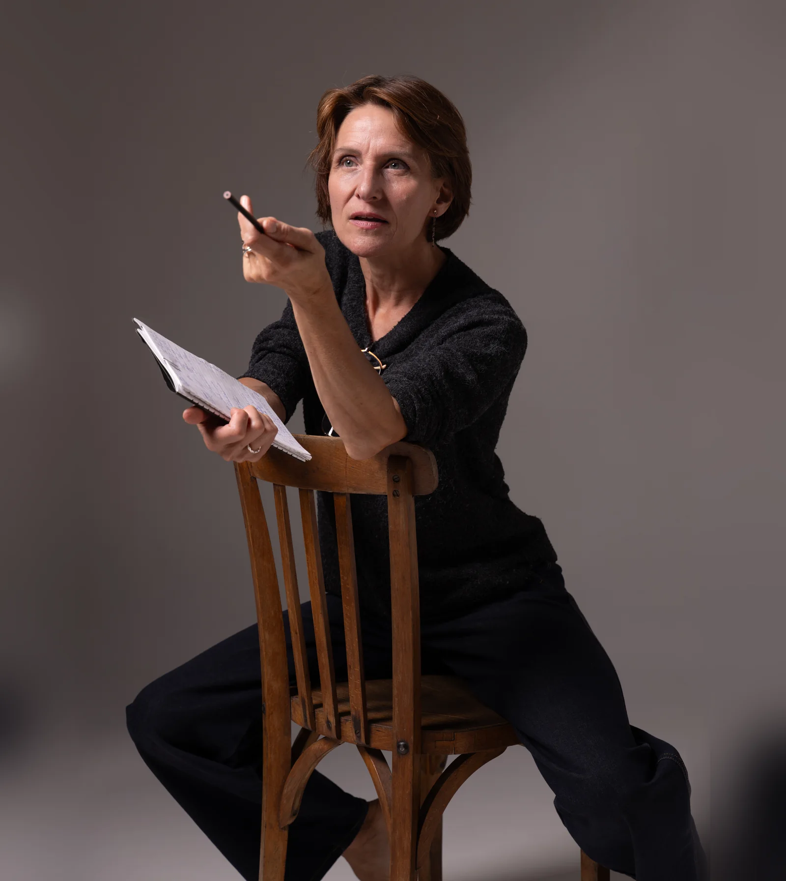 Laurence Arpi assise à califourchon sur une chaise de bois, carnet et crayon à la main, en séance de travail