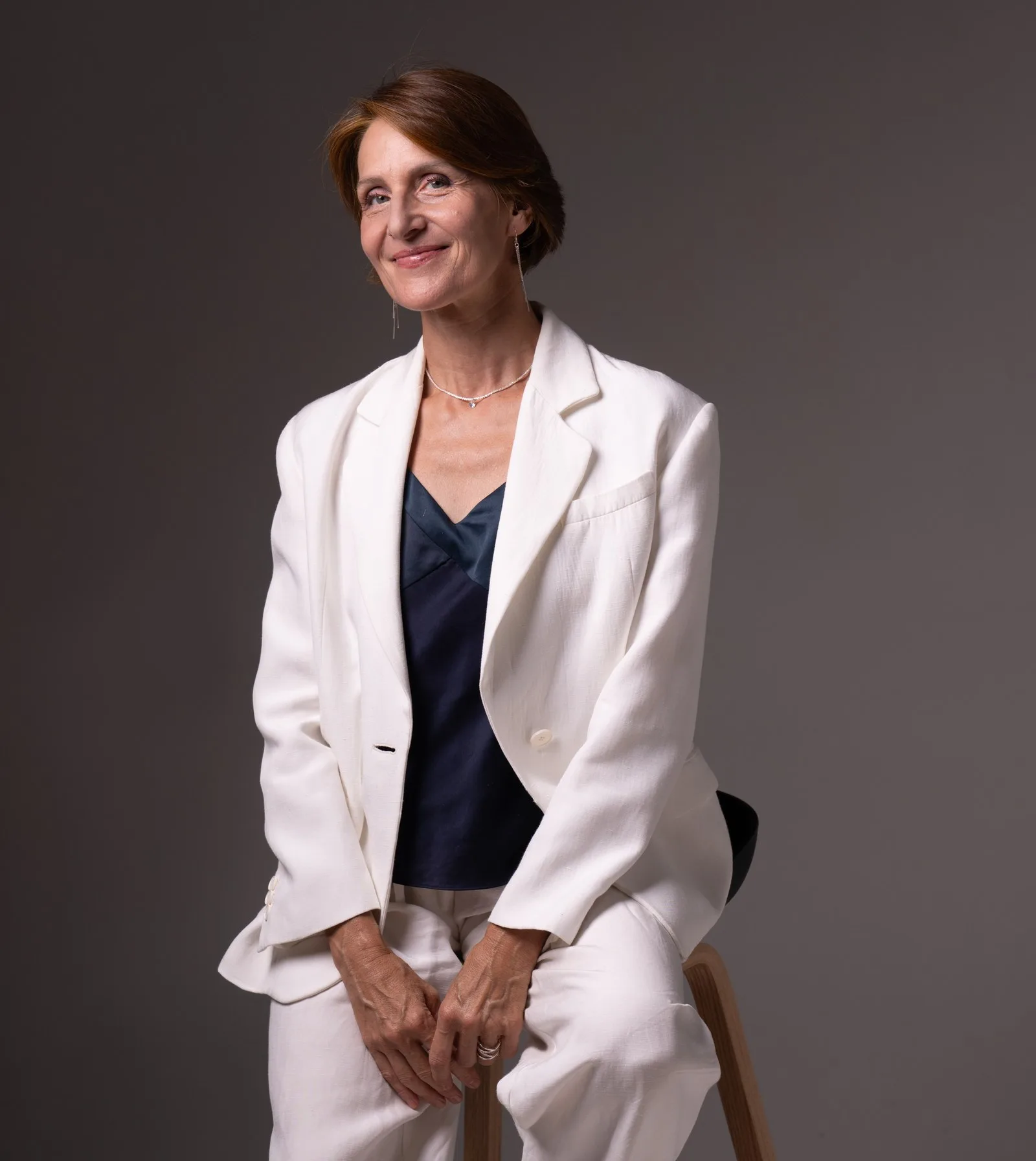 Portrait de Laurence Arpi assise sur un tabouret, costume blanc, sourire direct